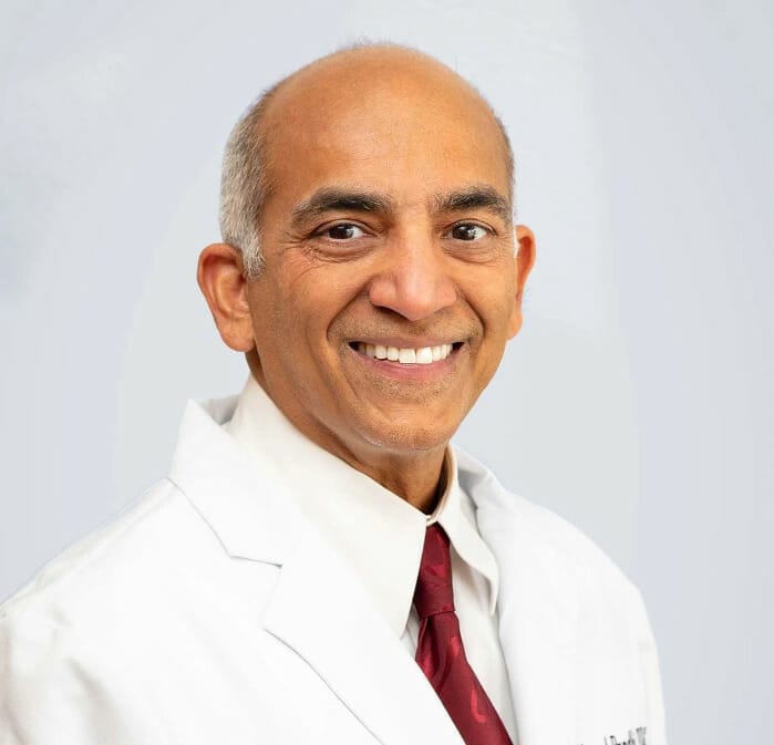 Dr. Pandhi smiling in a white coat and patterned tie, sits confidently in a professional medical office setting