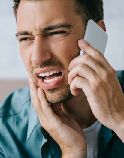 A man grimaces in pain while holding his jaw from TMJ and speaking on the phone, possibly experiencing a dental emergency