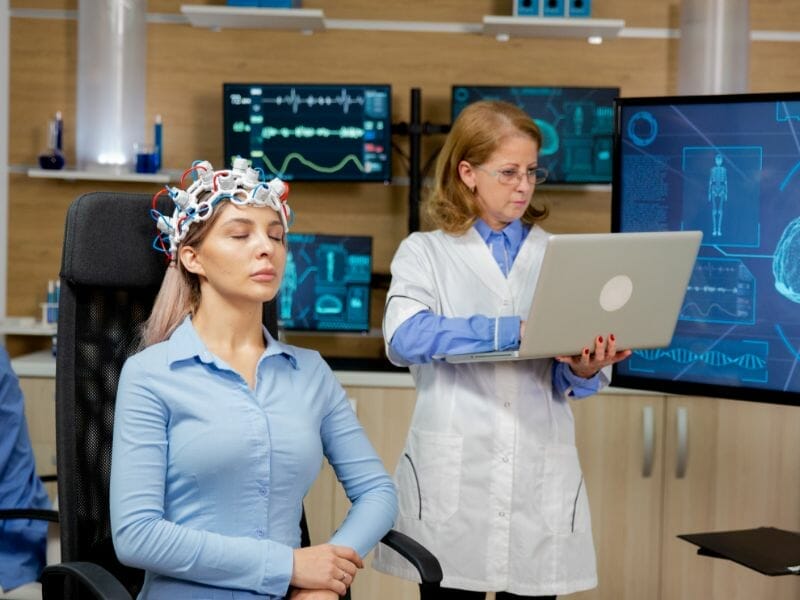 Patient undergoing a neurofeedback therapy session with brainwave monitoring cap, guided by a specialist analyzing real-time data on a laptop in a modern neuroscience lab.