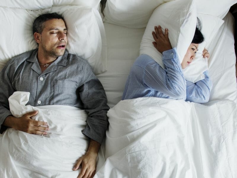 Man snoring in bed while his frustrated partner covers her ears with a pillow due to sleep disruption, illustrating the impact of snoring on sleep quality.
