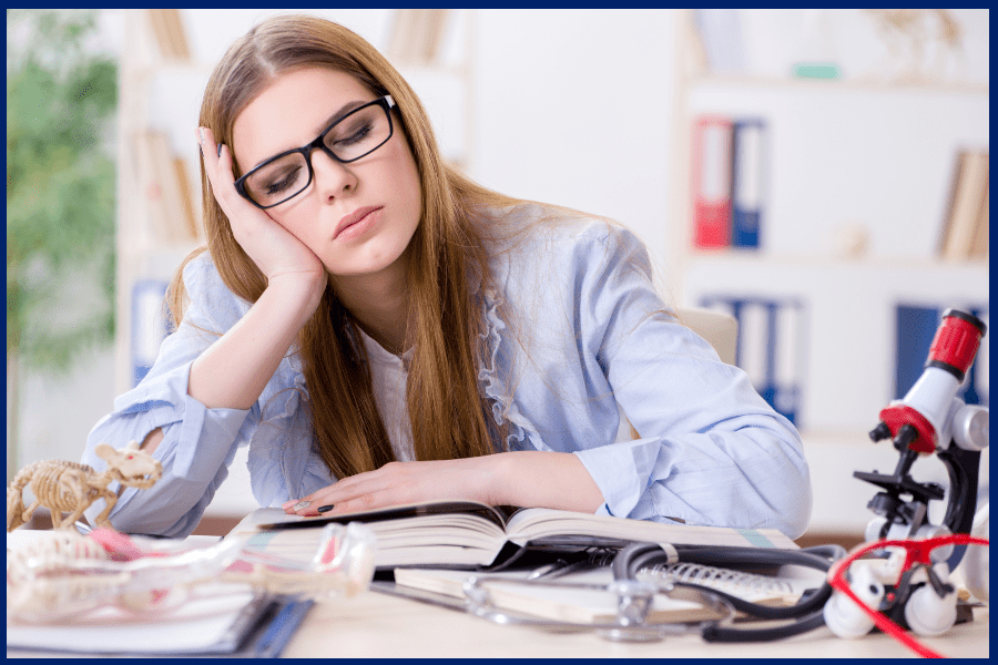 Female student experiencing study burnout, asleep over open textbooks with science tools like a stethoscope and microscope on the desk.