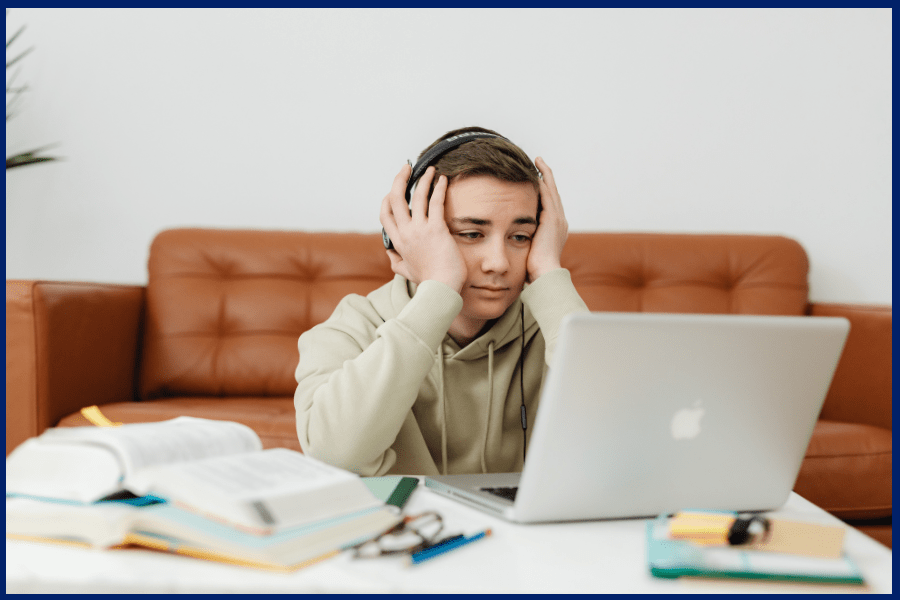Teen experiencing online learning fatigue, wearing headphones and looking tired in front of a laptop surrounded by study materials.