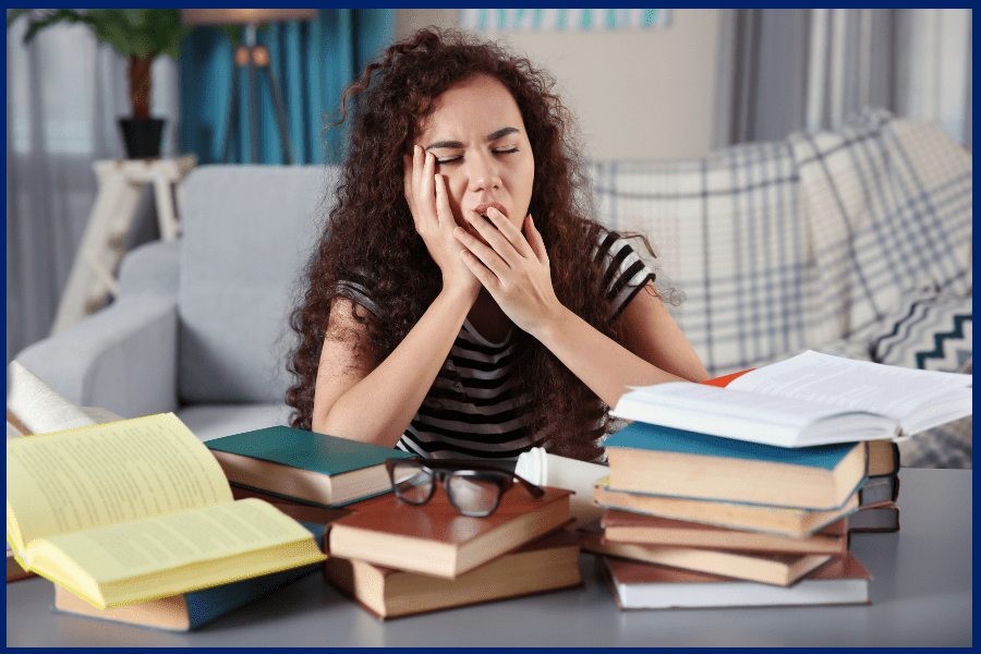 Exhausted student showing signs of study burnout, yawning at a desk surrounded by stacks of textbooks and notes at home.