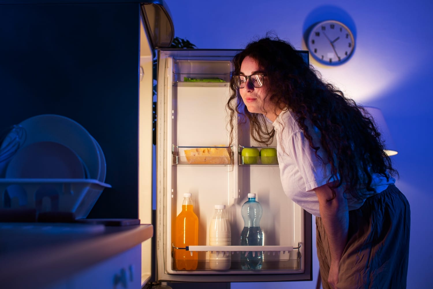Woman looking into an open refrigerator late at night, suggesting midnight snacking