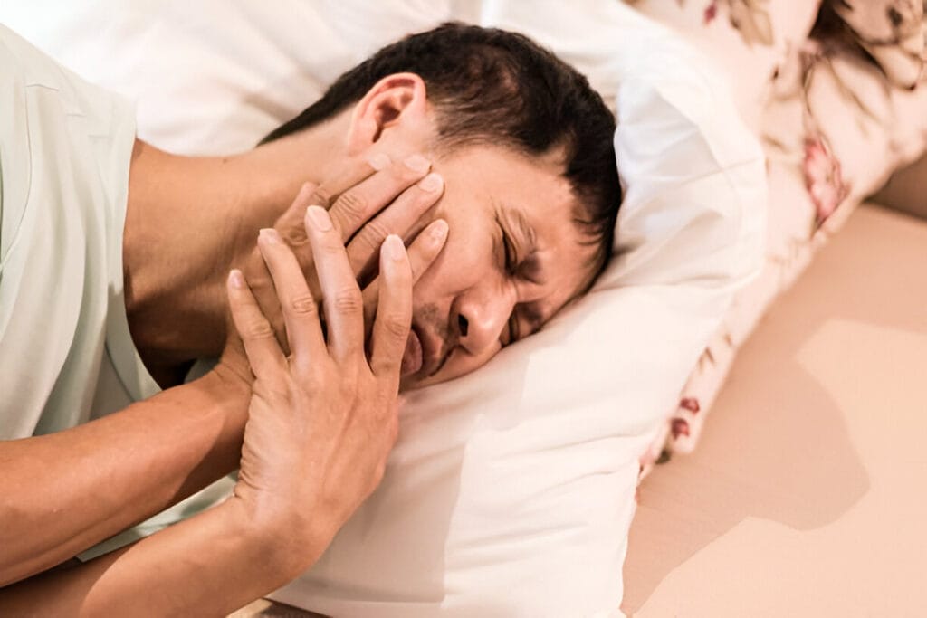 A man in bed clutching his jaw in pain, showing signs of TMJ discomfort