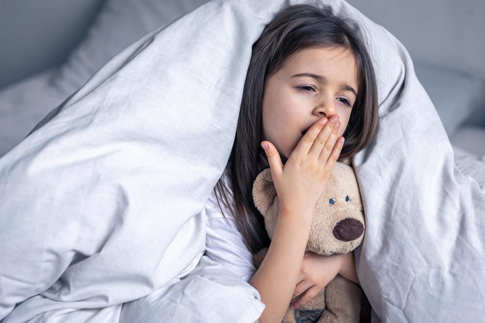 Young girl yawning under a blanket while holding a stuffed bear