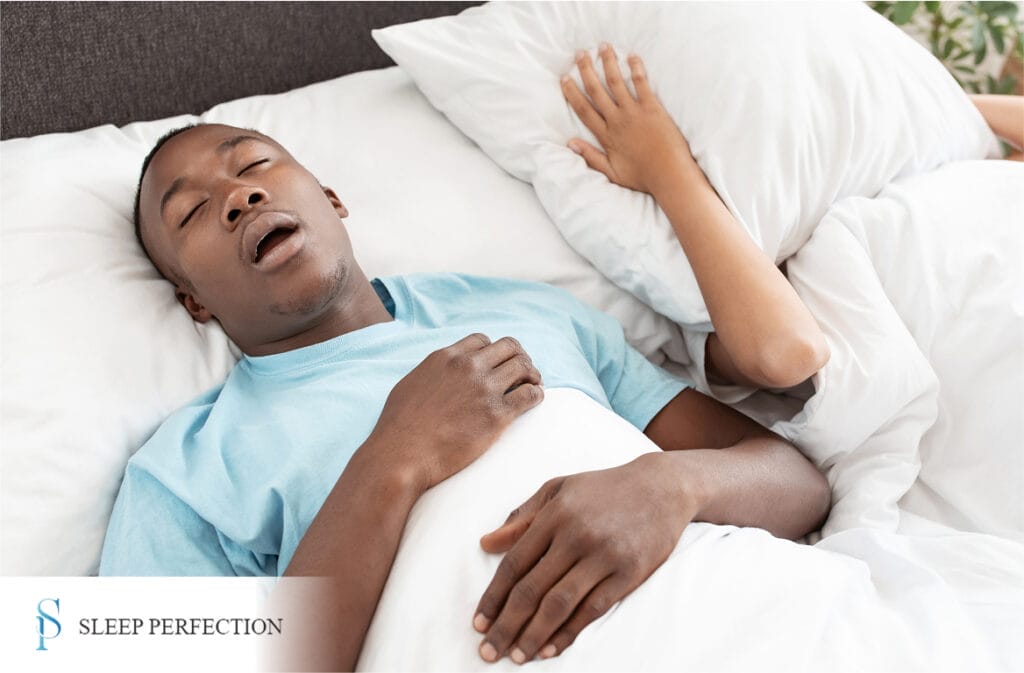 A man sleeps with his mouth open while a partner next to him covers their ears with a pillow